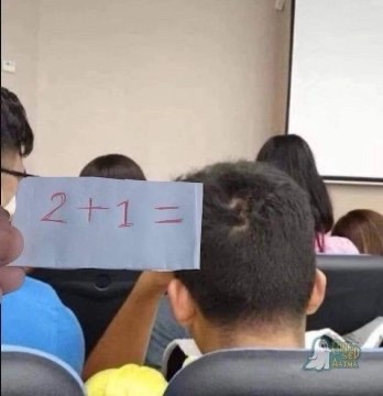 A person in a classroom holds up a small piece of paper with the math problem '2+1=' written in red marker. The paper is…