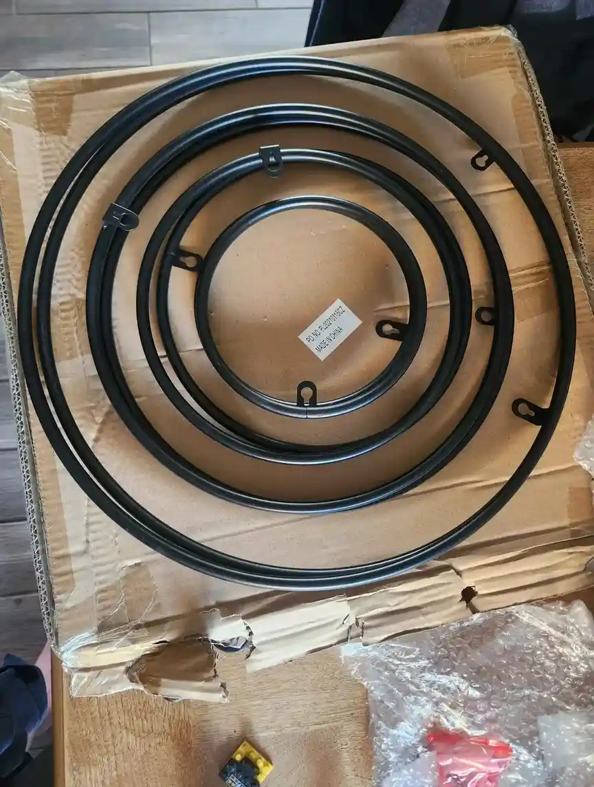 An overhead photo shows three concentric black metal rings, each with small hooks, resting on a cardboard box. A small white…