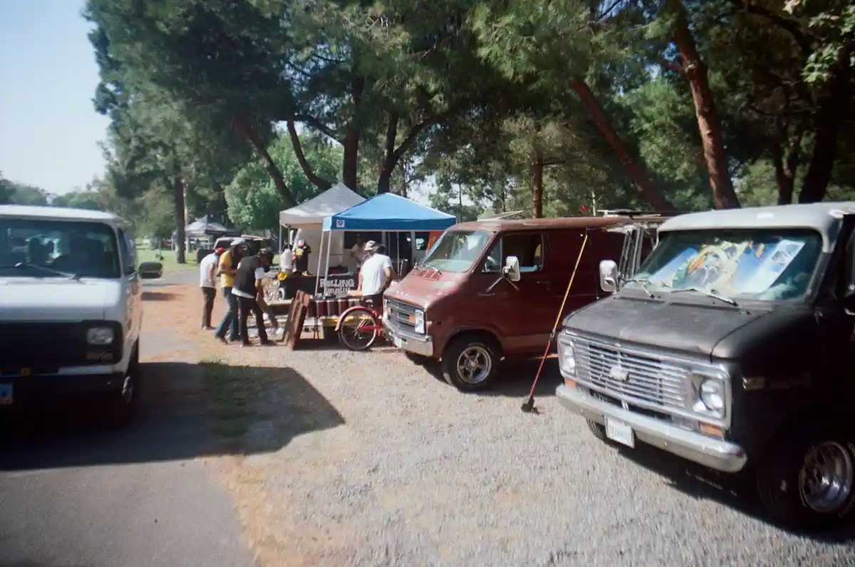 A vintage photo shows several classic vans parked on gravel under trees at an outdoor gathering. People are gathered around…