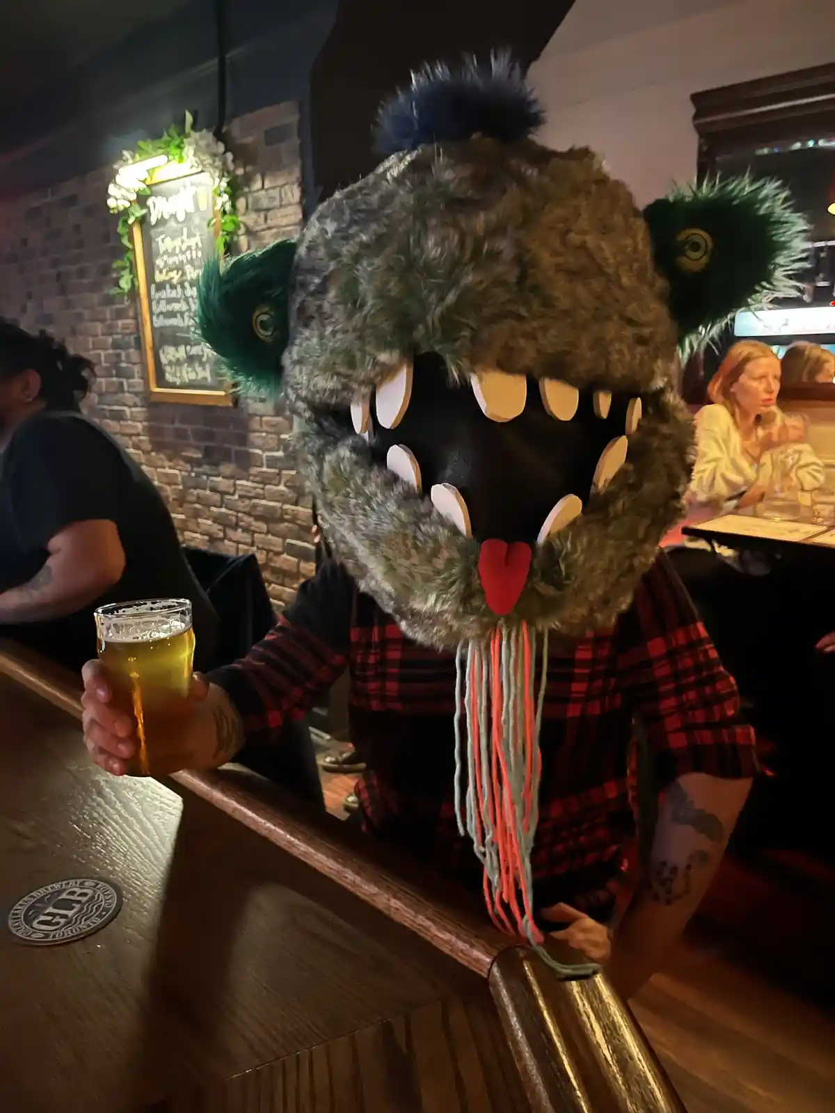 A person wearing a large, furry, toothy monster mask with green ears and a red tongue is holding a glass of beer at a bar…