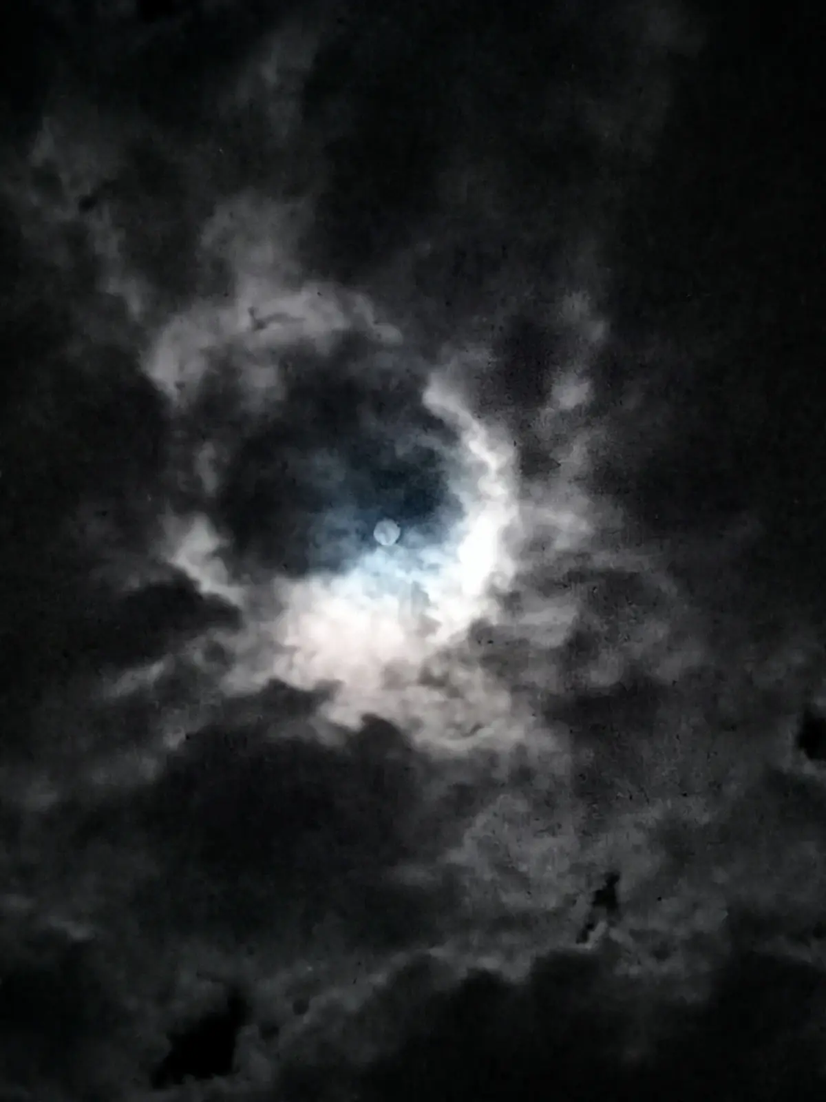 A dark, moody night sky with thick clouds partially obscuring a bright, glowing moon. The image has a grainy, low-light…