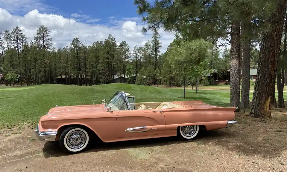 A vintage peach-colored convertible car with chrome details and whitewall tires is parked on dirt near pine trees, with a…