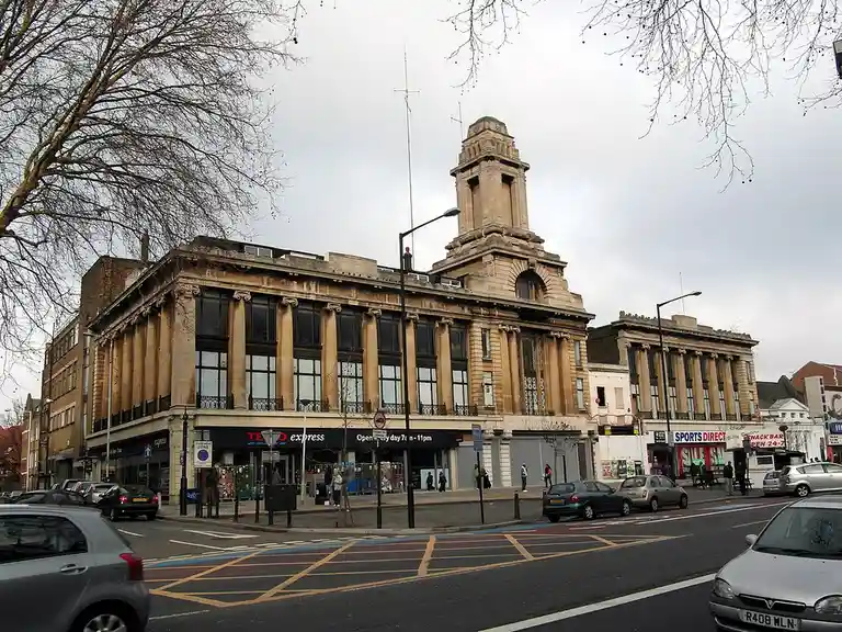 A street-level photo of a large, ornate, classical-style building with a clock tower, now housing a 'Tesco Express'…