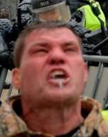 A close-up photo of a man with a furious, yelling expression, his mouth wide open showing teeth, and a small white object…