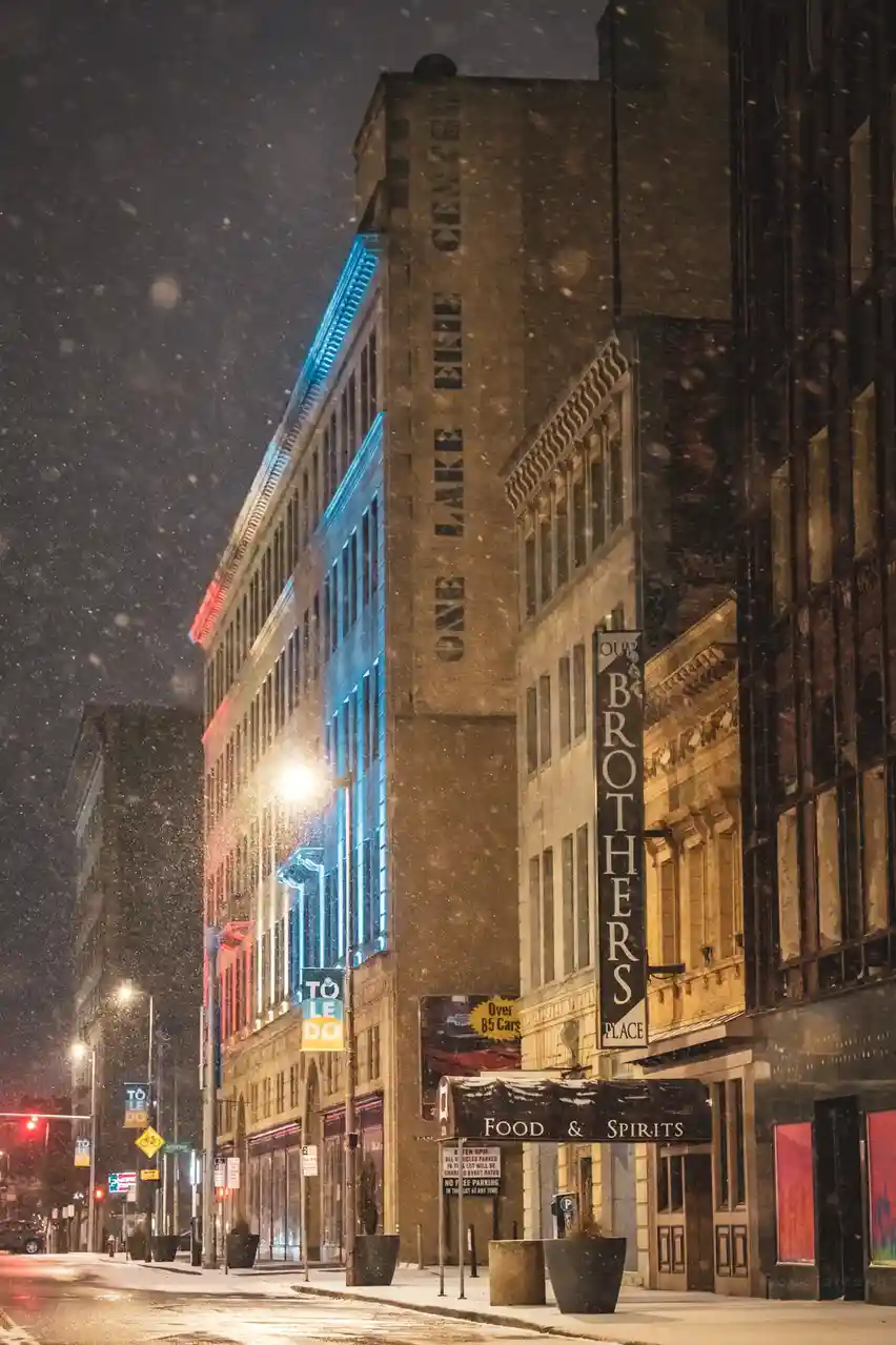 A snowy night city street with illuminated buildings, one prominently lit in blue and red, featuring a vertical sign that…