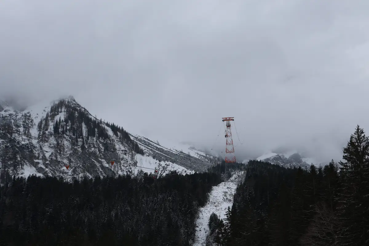 A snowy mountain landscape with evergreen trees and a red-and-white cable car tower partially obscured by clouds. Several…
