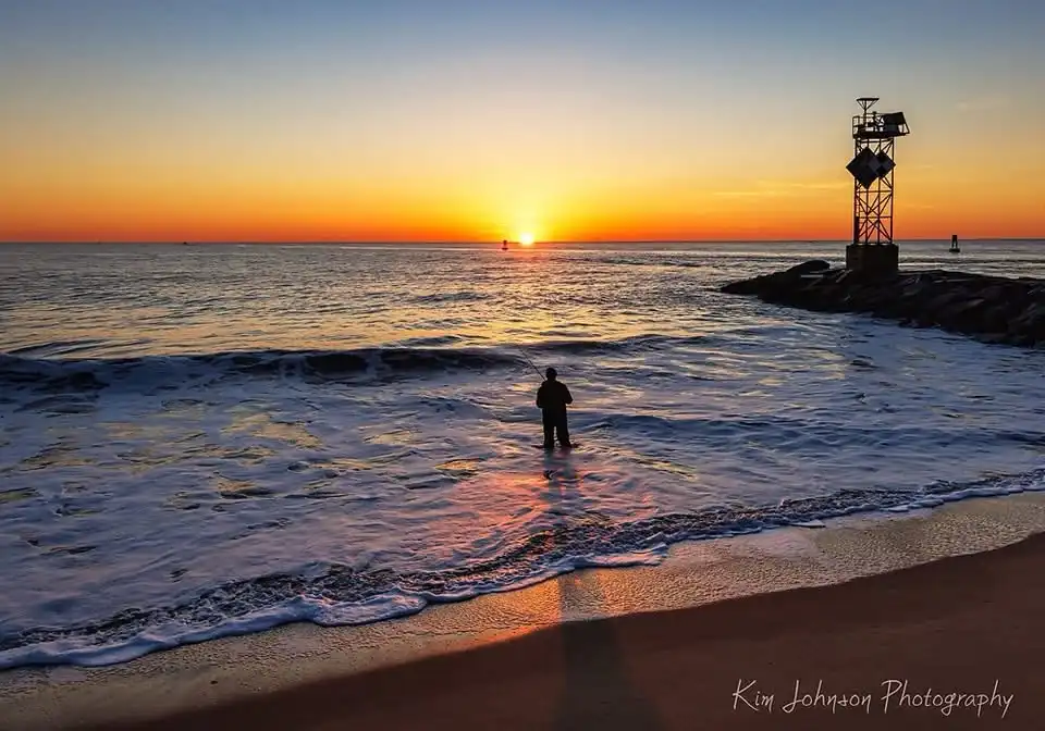 A silhouette of a person standing knee-deep in ocean water at sunset, holding a fishing rod. The sky is painted with orange…
