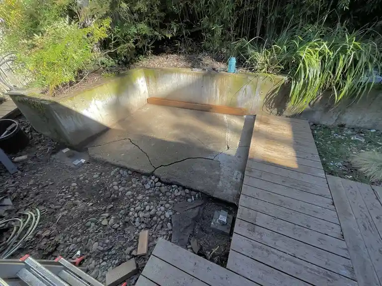 An outdoor concrete structure resembling a hot tub, cracked and unfinished, sits beside a wooden deck with tools and debris…