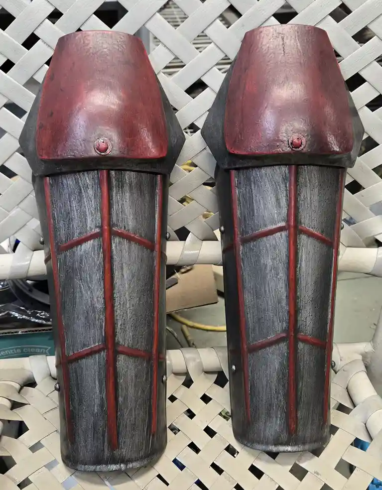 A pair of custom-made, red and gray fantasy-style shin guards with a wooden texture and red trim, resting on a white lattice…