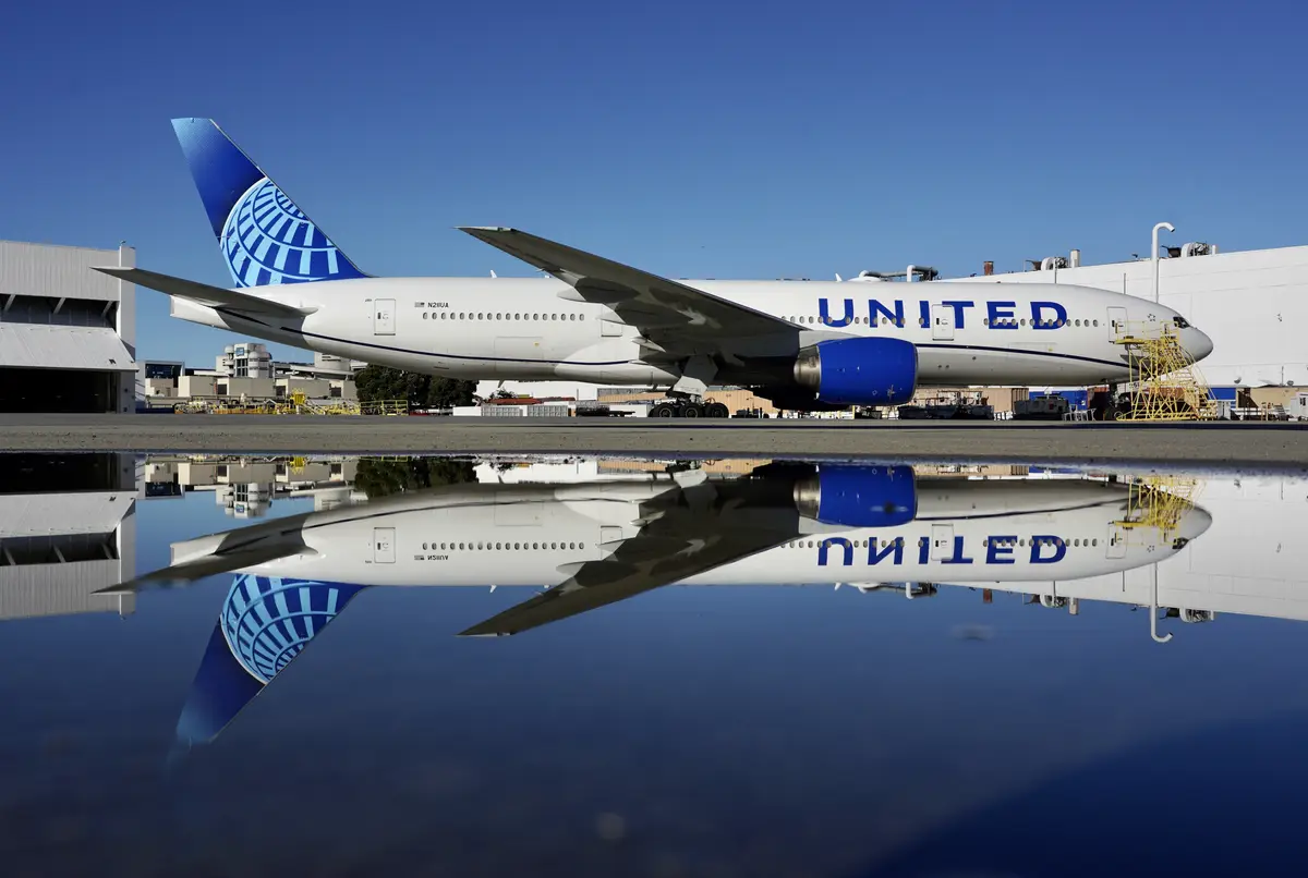 A United Airlines passenger jet is parked on an airport tarmac under a clear blue sky, with its reflection perfectly…