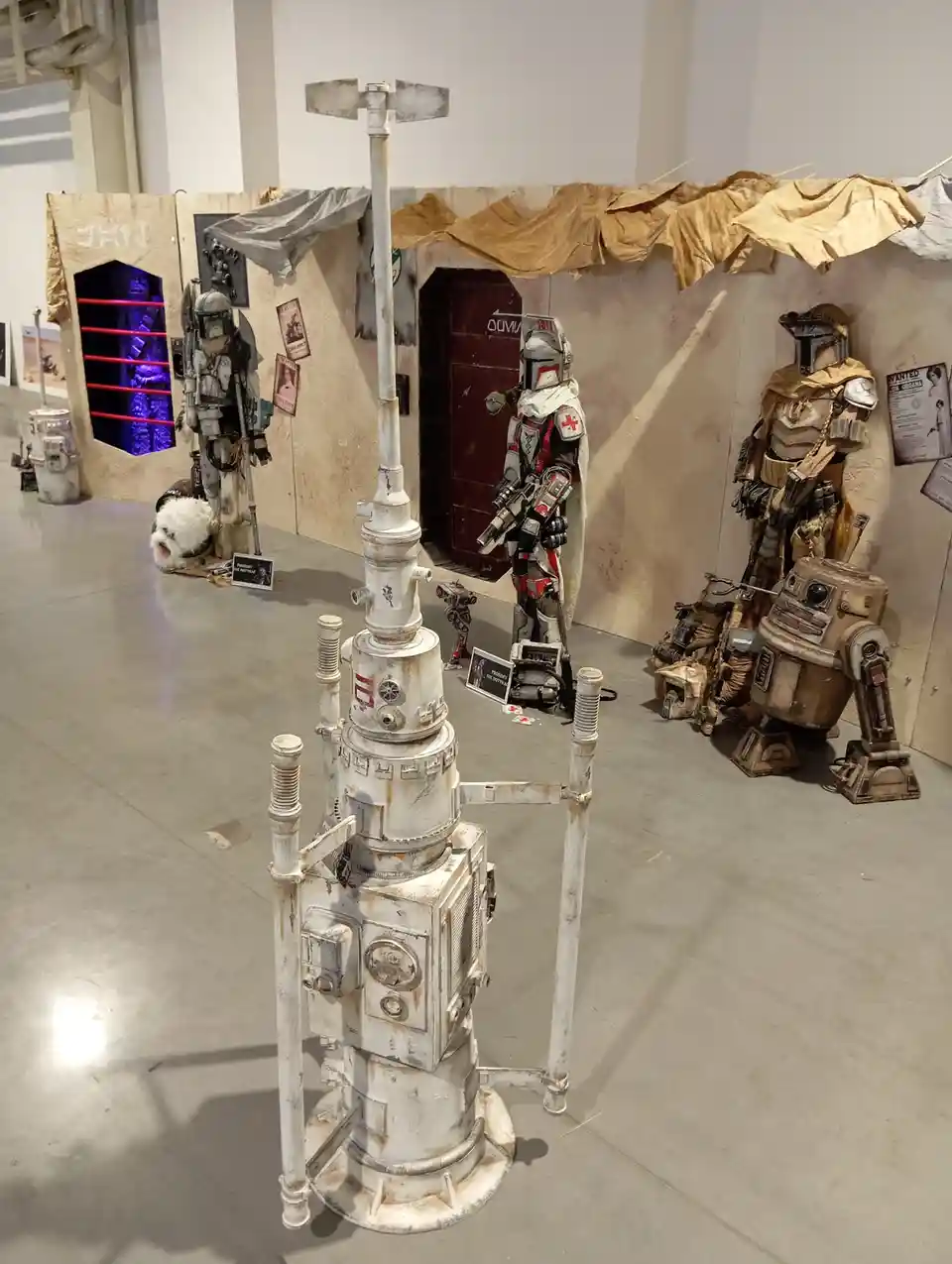 A photo of a Star Wars-themed cosplay exhibit featuring Mandalorian armor, a droid, and a small white dog in a costume, with…