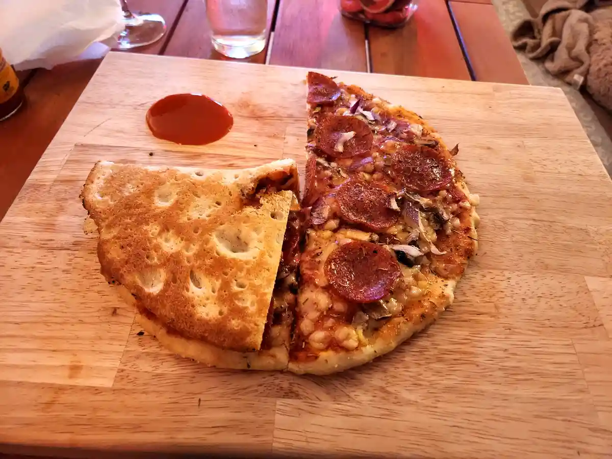 A slice of pepperoni pizza sits on a wooden board next to a folded piece of toasted bread, with a dollop of red sauce…