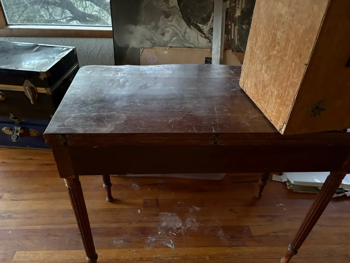 A worn wooden table with a scratched surface and a wooden box leaning against it, set on a hardwood floor with dust and…