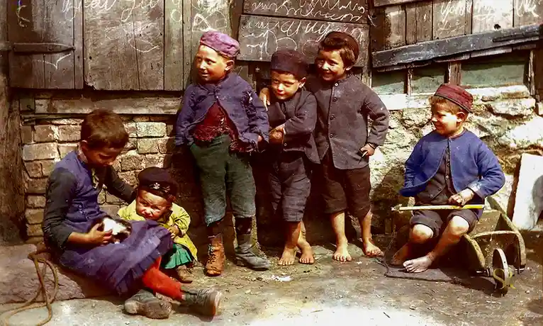 A vintage colorized photo shows seven young boys in worn clothing, some barefoot, gathered outside a rustic wooden building…