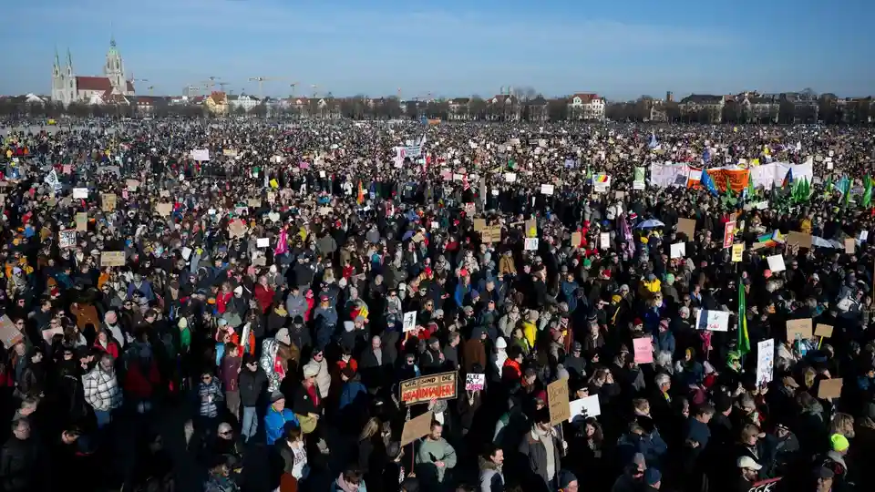 A vast crowd of people fills a large public square under a clear blue sky, holding numerous handmade signs and banners. In…