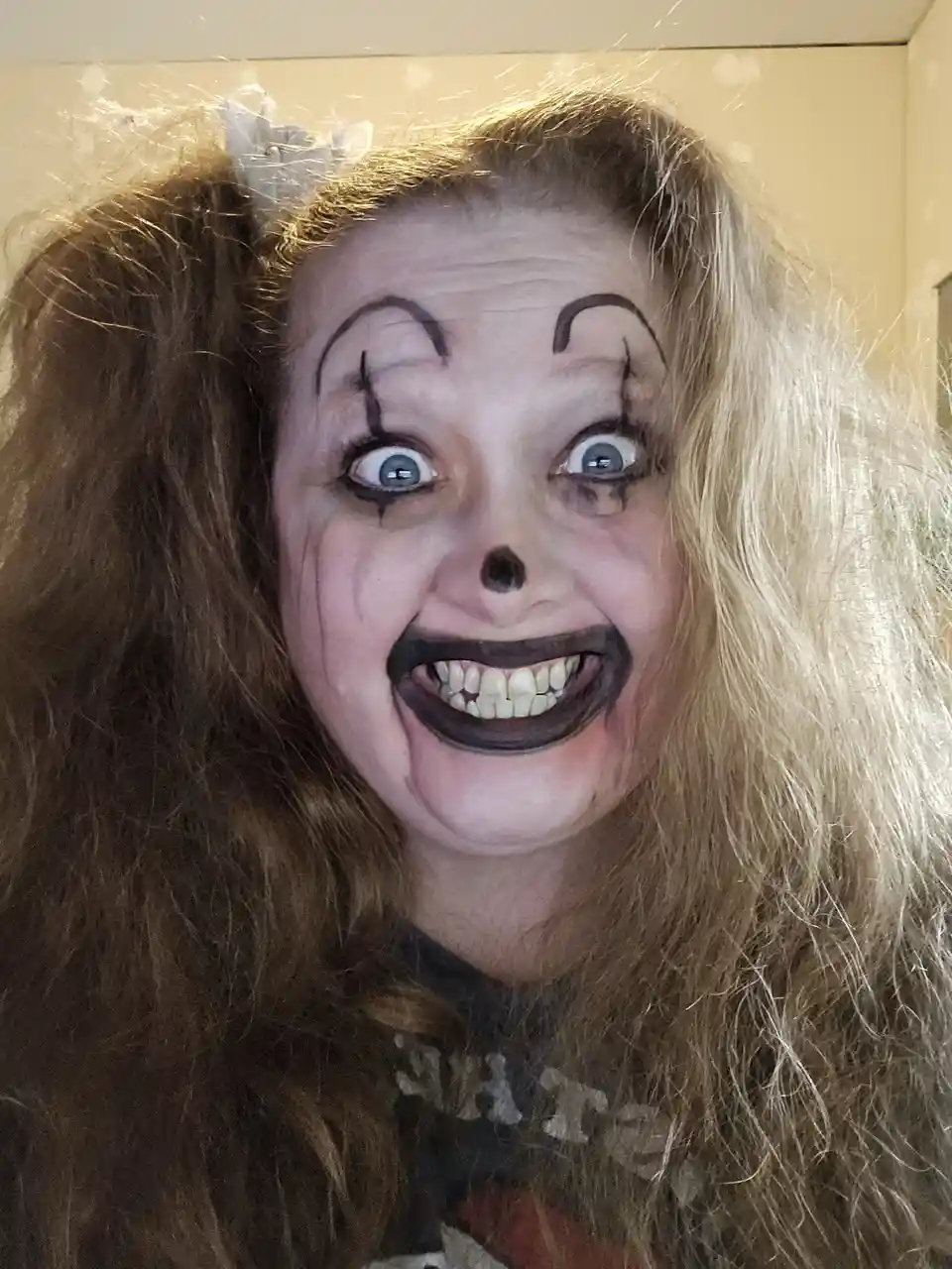 A person with long, messy blonde hair wears exaggerated clown makeup featuring white face paint, black arched eyebrows…