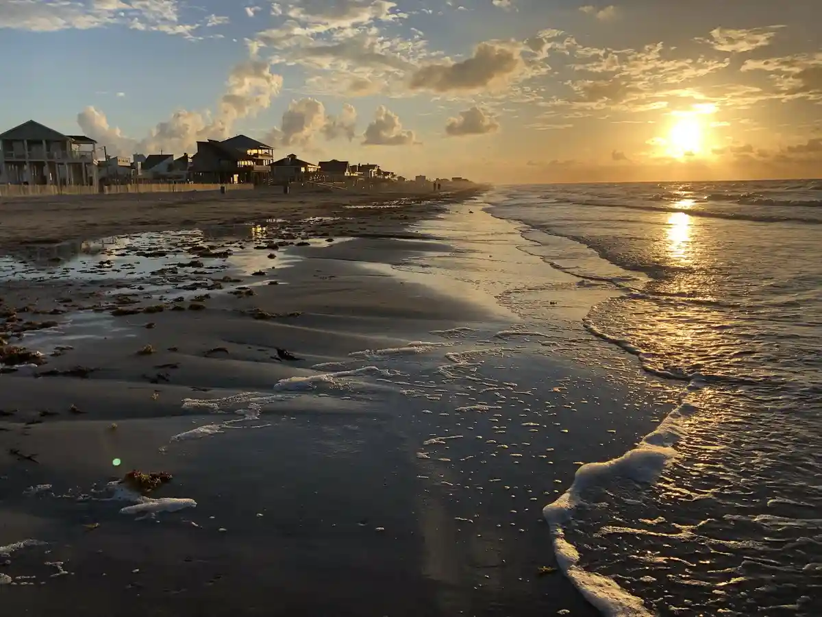 A serene beach scene at sunset with waves lapping at the shore, scattered seaweed, and beachfront houses in the distance…