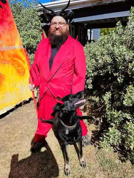 A man in a red devil costume with horns walks a black dog outdoors in front of a fiery backdrop. The dog’s expression…