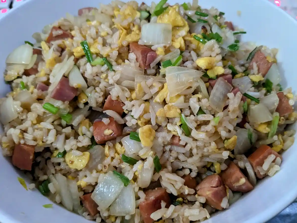 a close-up photo of a white bowl filled with fried rice mixed with scrambled egg, chopped green onions, diced onions, and…