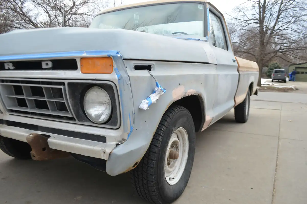 A vintage Ford truck is shown in the middle of a paint job, with blue painter's tape covering parts of the body and a…