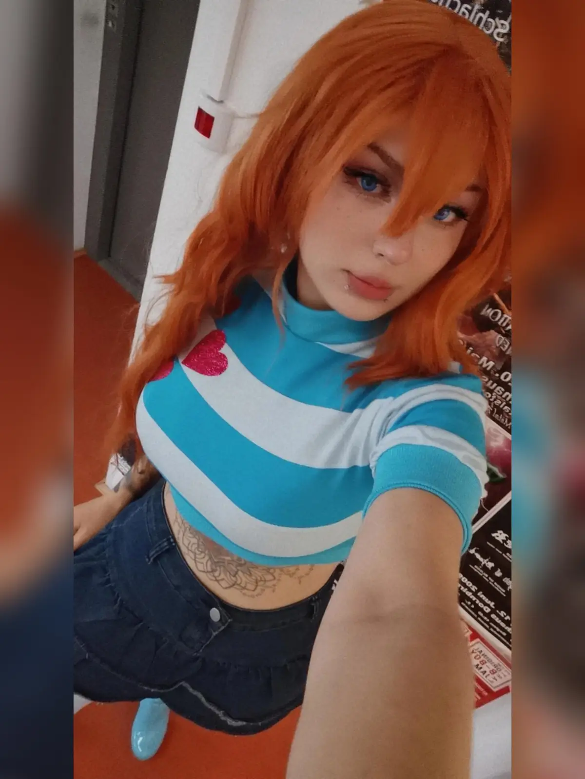 A person with bright orange hair and blue eyes takes a selfie wearing a blue and white striped crop top with a pink glitter…