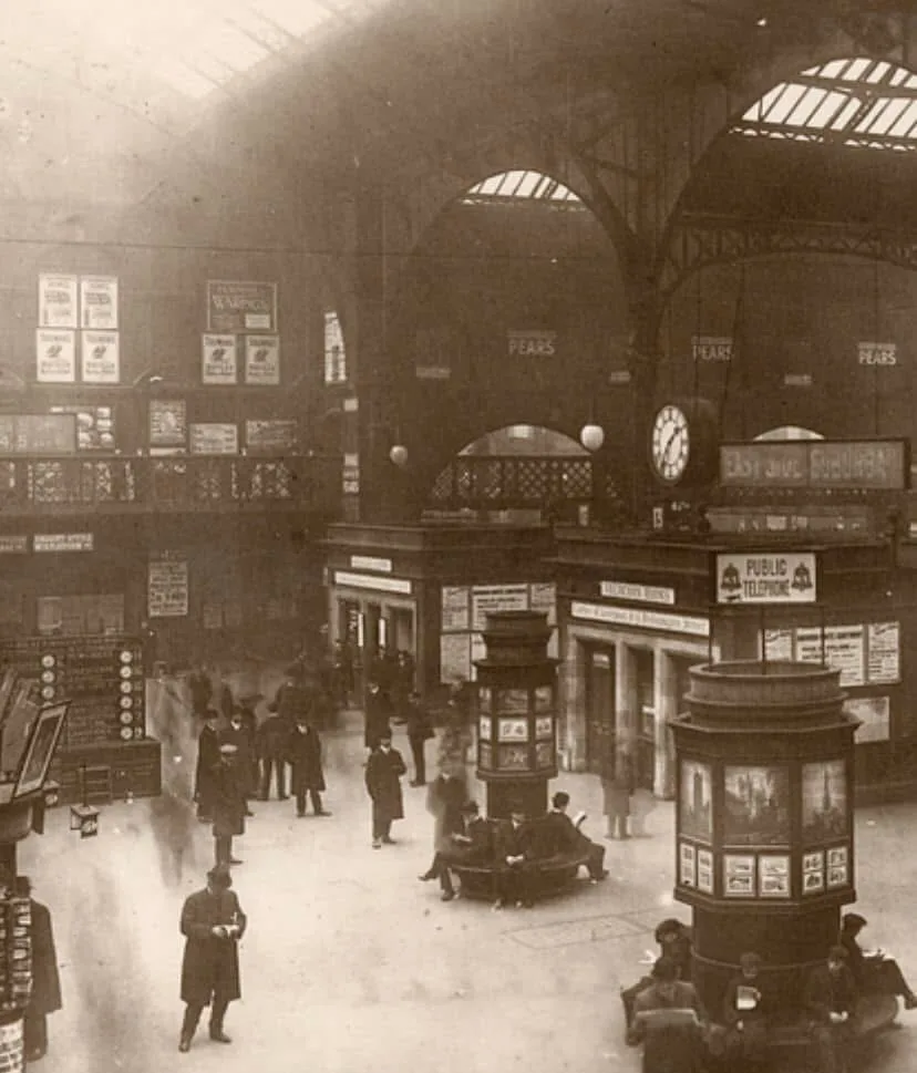 A sepia-toned historical photograph of a bustling, high-ceilinged train station from the early 20th century, filled with…