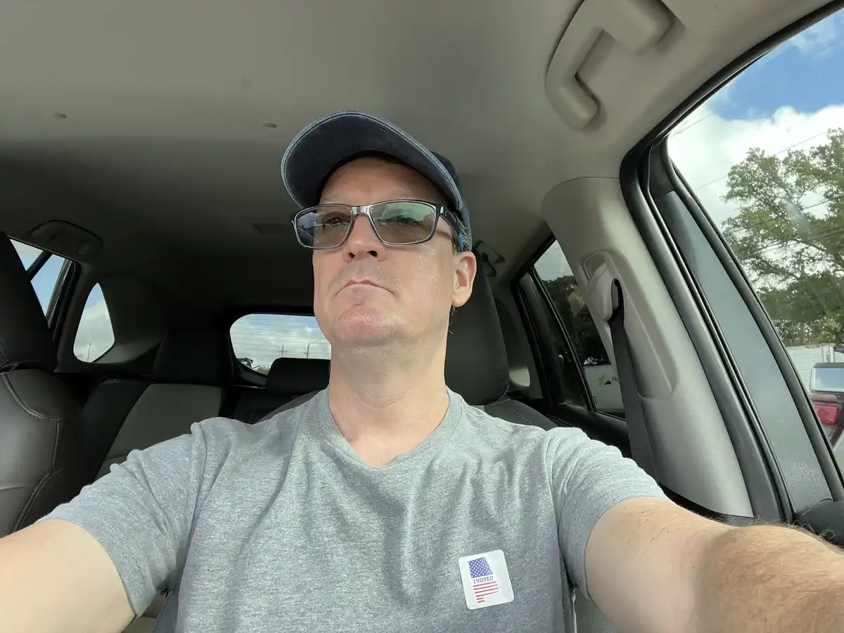 A man wearing a grey t-shirt with an 'I VOTED' sticker, a baseball cap, and glasses takes a selfie inside a car. He has a…