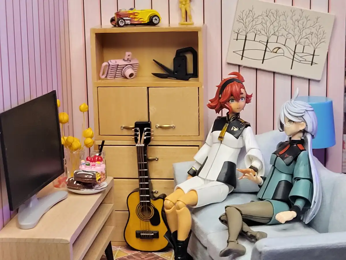 Two anime-style figurines sit on a miniature couch in a dollhouse-like room with pink walls, a wooden cabinet, a guitar, and…