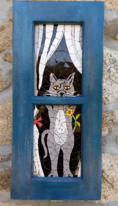 A mosaic artwork on a blue wooden window frame depicts a cat with a human-like face peering through the top pane, while the…