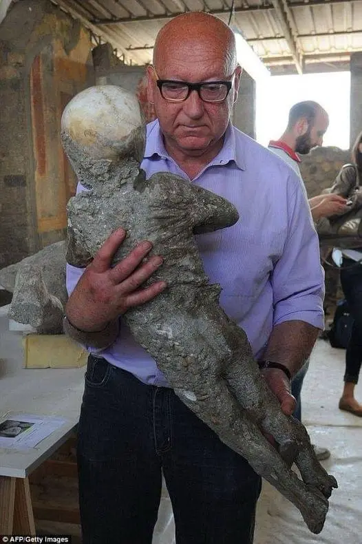 A bald man with glasses holds a small, gray, clay-like statue of a child. The statue has a round head and is held gently…