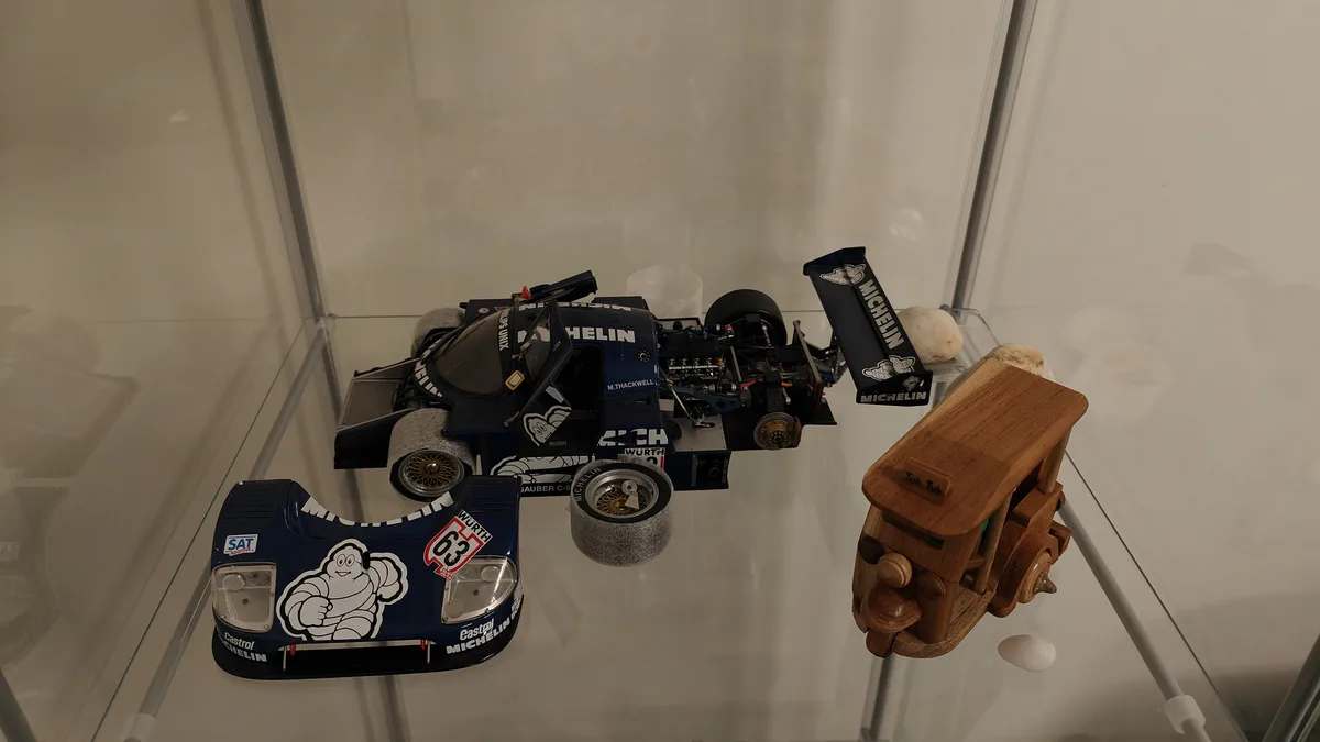 A glass display case contains a detailed blue and white scale model of a Michelin-sponsored racing car with visible engine…