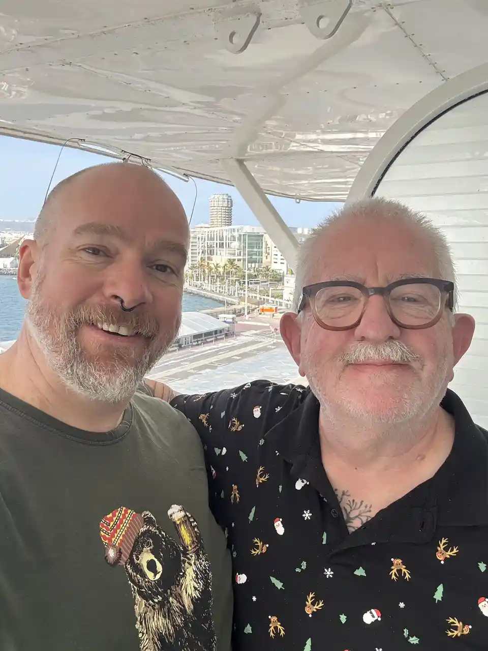 Two men are taking a selfie inside what appears to be a small aircraft or boat cabin, with a city waterfront visible through…