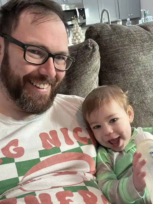 A smiling man with glasses and a beard sits on a couch holding a baby who is also smiling and holding a bottle. The baby has…