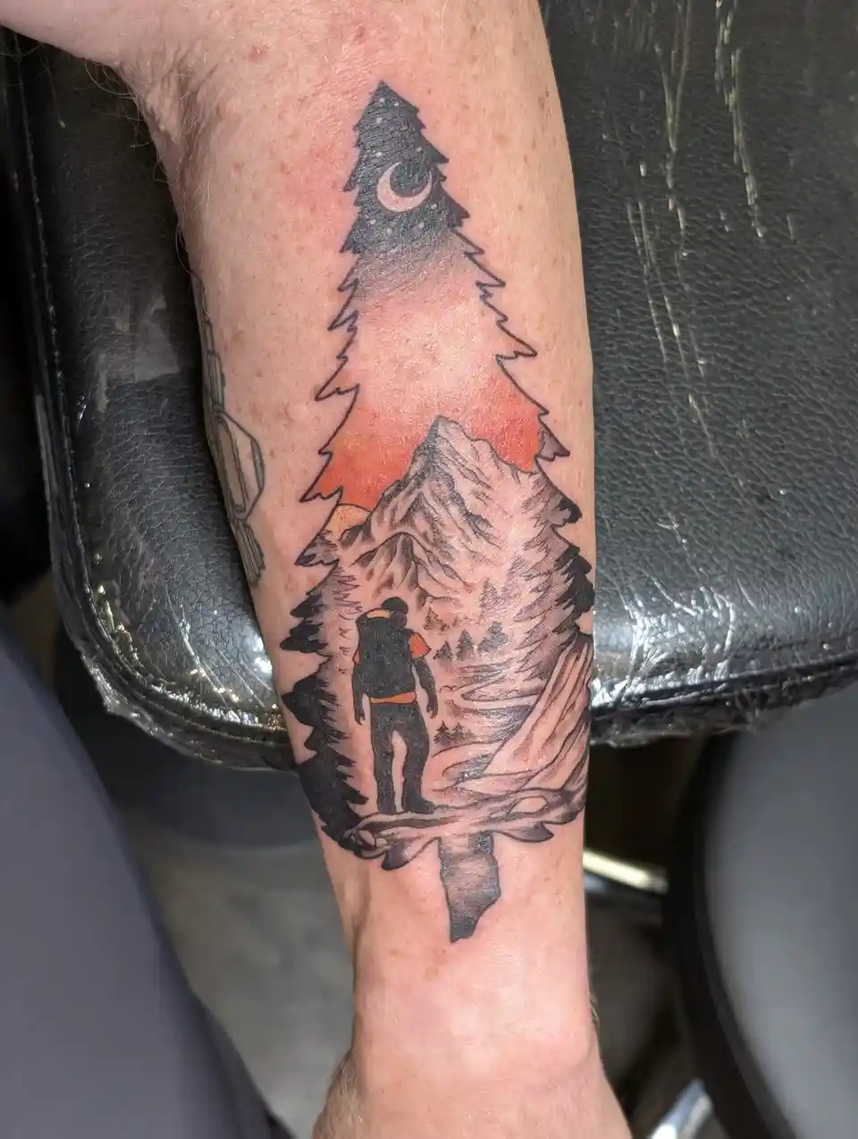 A close-up photo shows a tattoo on a person's forearm depicting a hiker standing on a path, looking up at a large pine tree…
