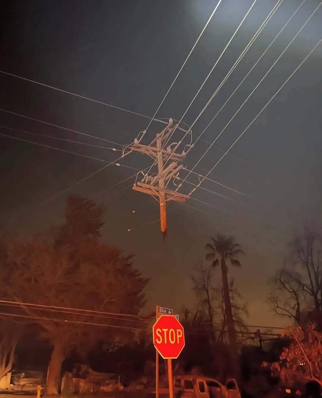 A nighttime photo shows a wooden utility pole hanging precariously by wires, suspended in mid-air above a street with a…