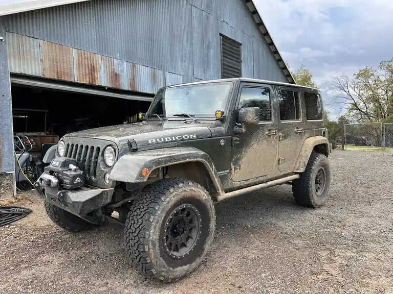 A heavily mud-splattered dark green Jeep Wrangler Rubicon is parked on gravel in front of a rusty corrugated metal barn with…