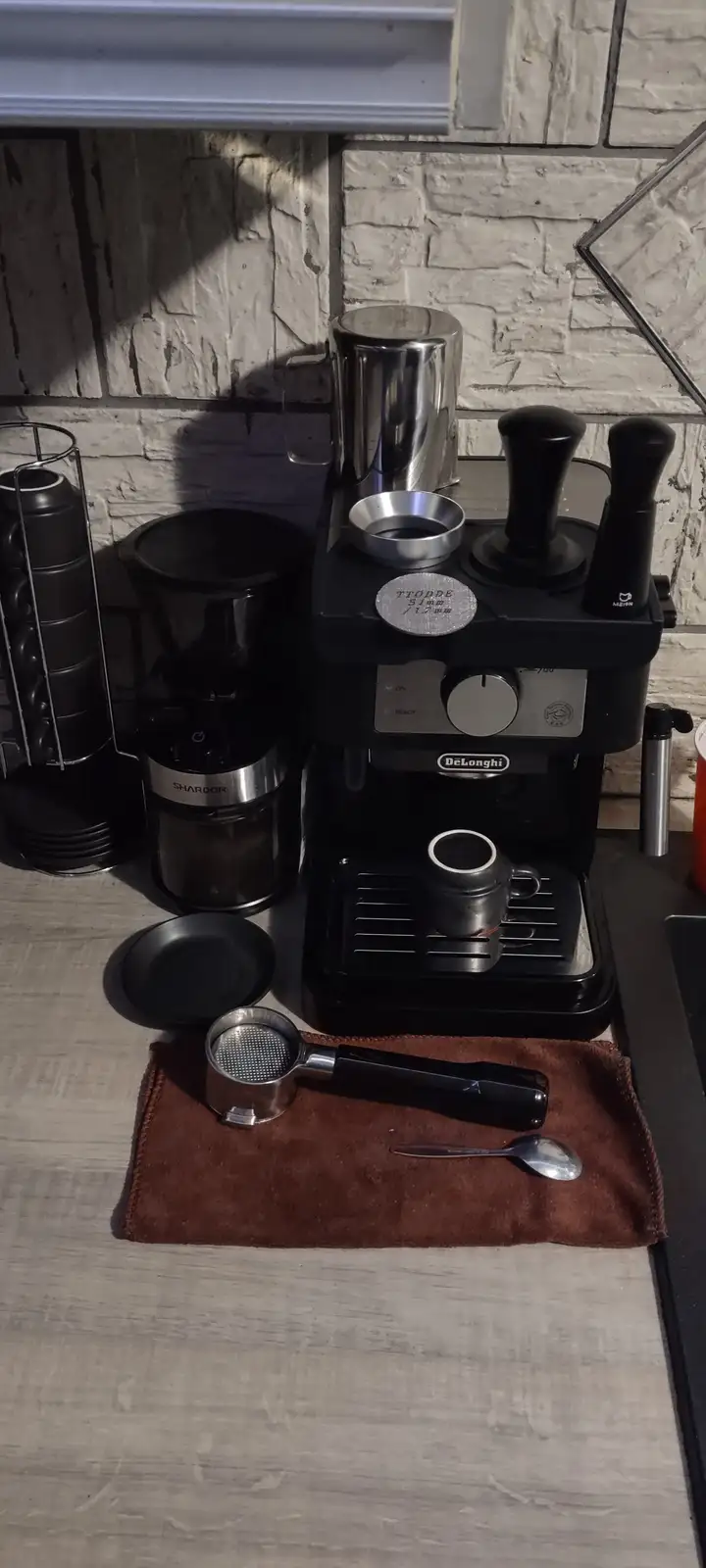 A black DeLonghi espresso machine sits on a kitchen counter with various coffee-making tools around it, including a…