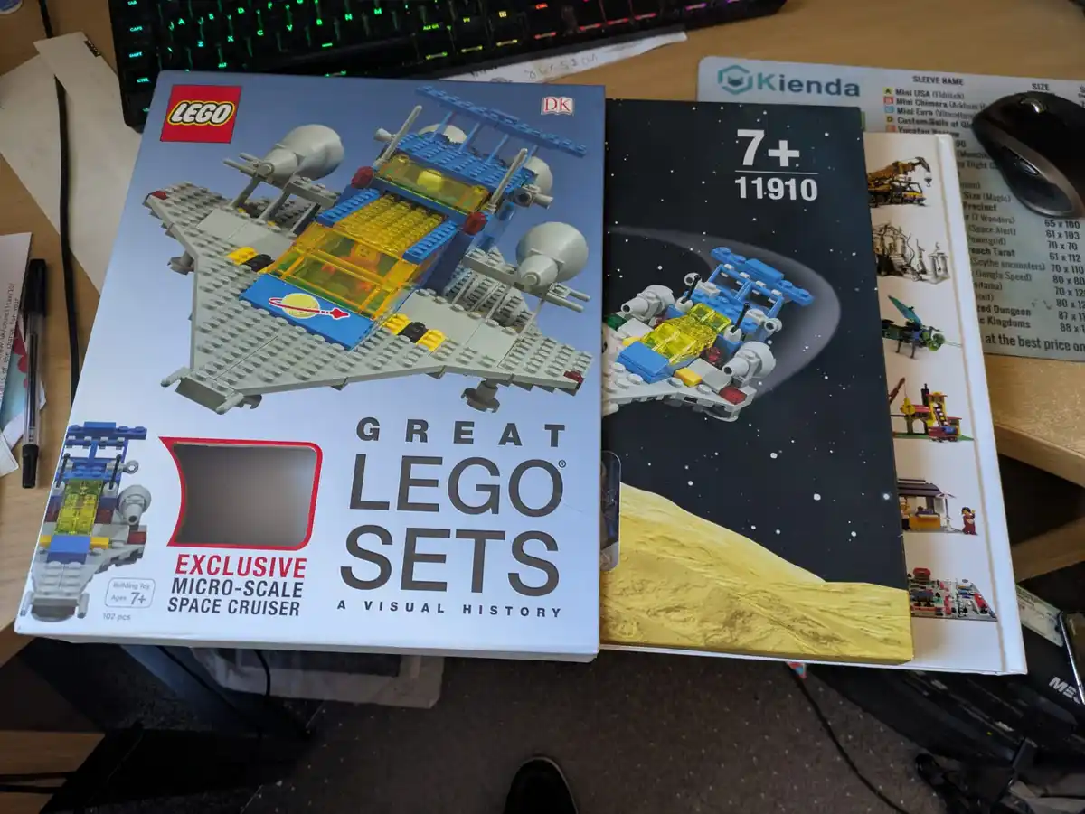 Two books are shown side-by-side on a desk: one is 'Great LEGO Sets: A Visual History' featuring a LEGO micro-scale space…