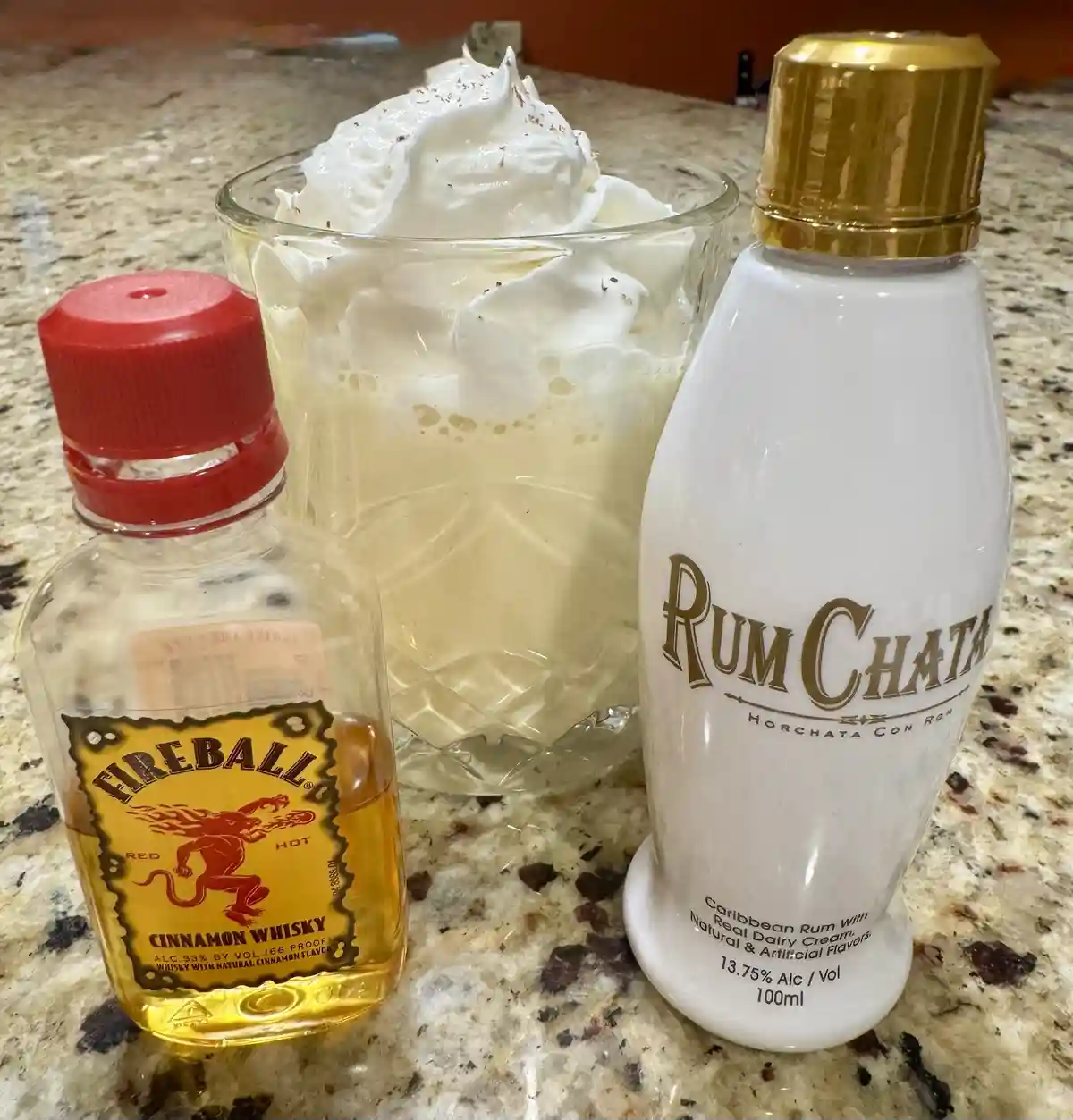 A glass of creamy, whipped-topped drink sits between a small bottle of Fireball Cinnamon Whisky and a white bottle of…