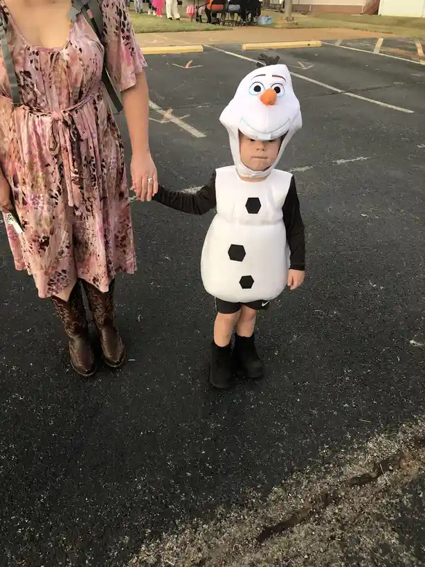 A young child wearing a full Olaf costume from Frozen, complete with a hooded headpiece and a white body with black hexagon…