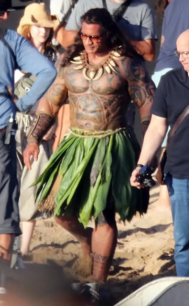 A man with long dark hair, sunglasses, and extensive tribal tattoos is wearing a green leaf skirt and a bone necklace while…