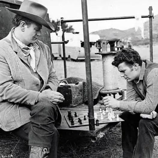 Two men in cowboy attire are intensely focused on a game of chess outdoors. The man on the left, wearing a wide-brimmed hat…