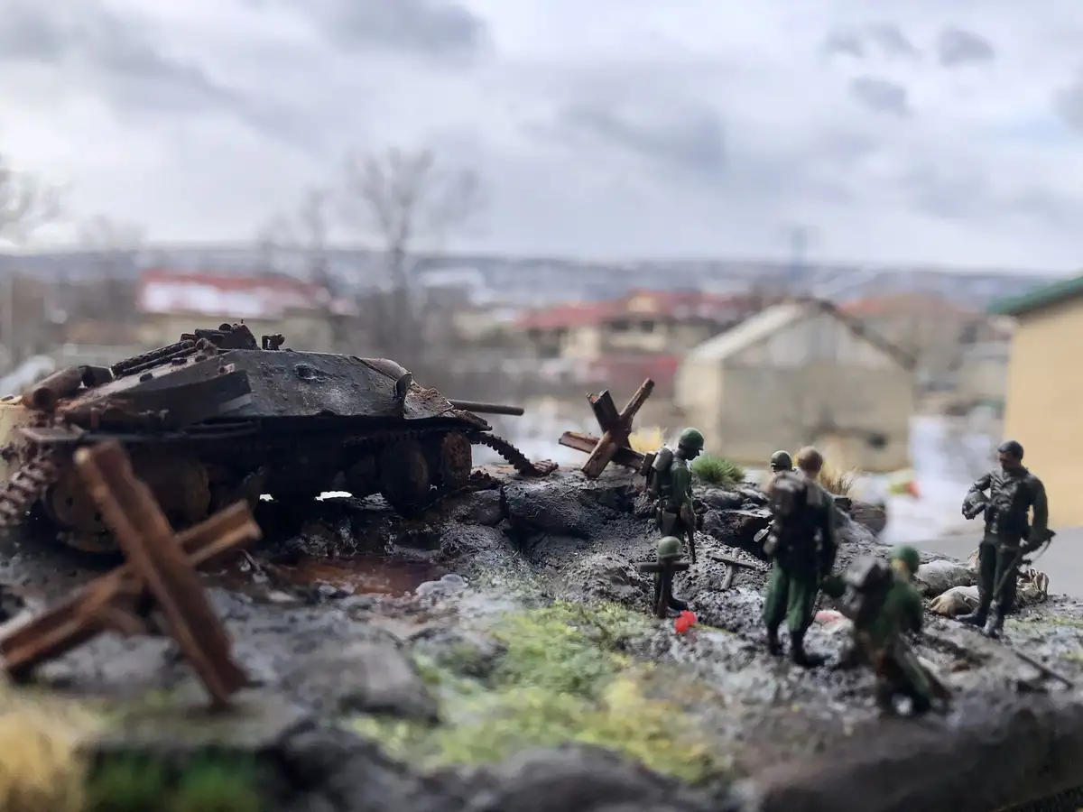 A close-up photo of a miniature diorama featuring a rusted toy tank and several toy soldiers standing on a muddy, rocky…