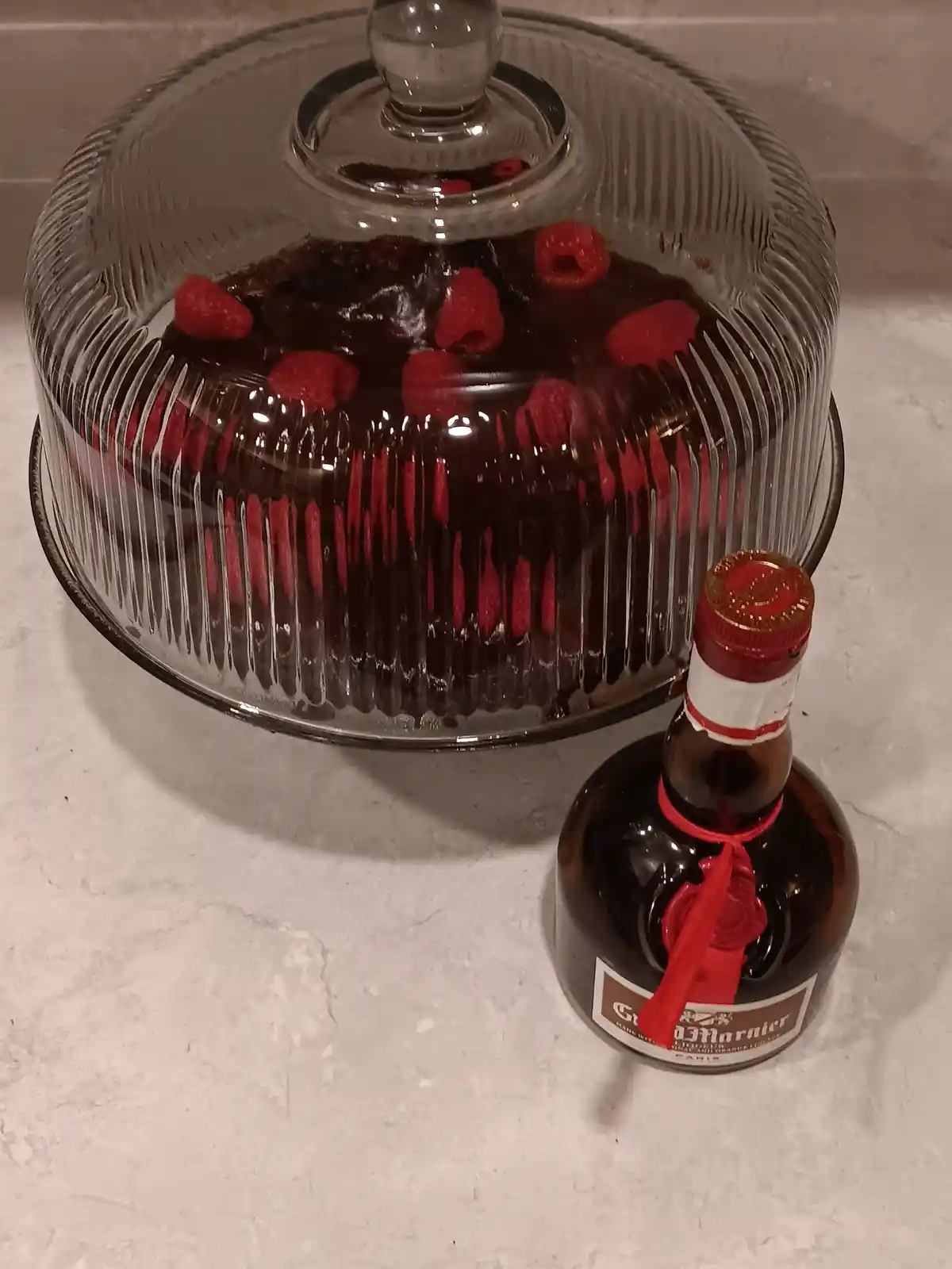 A chocolate cake topped with raspberries sits under a glass dome on a white countertop, next to a bottle of Grand Marnier…
