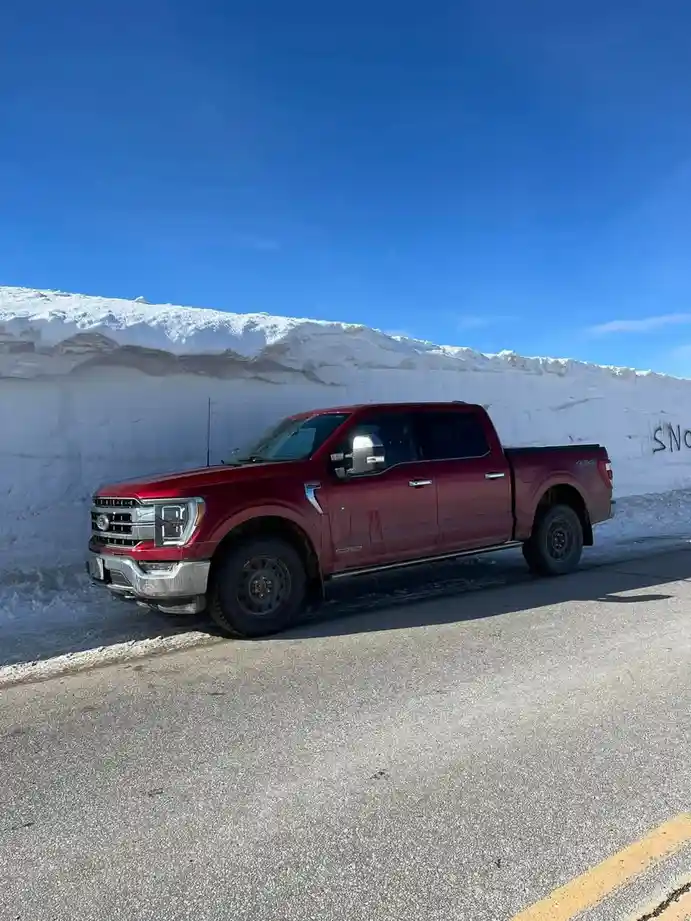 A red Ford F-150 pickup truck is parked on an asphalt road next to a tall, man-made snow wall under a clear blue sky. The…