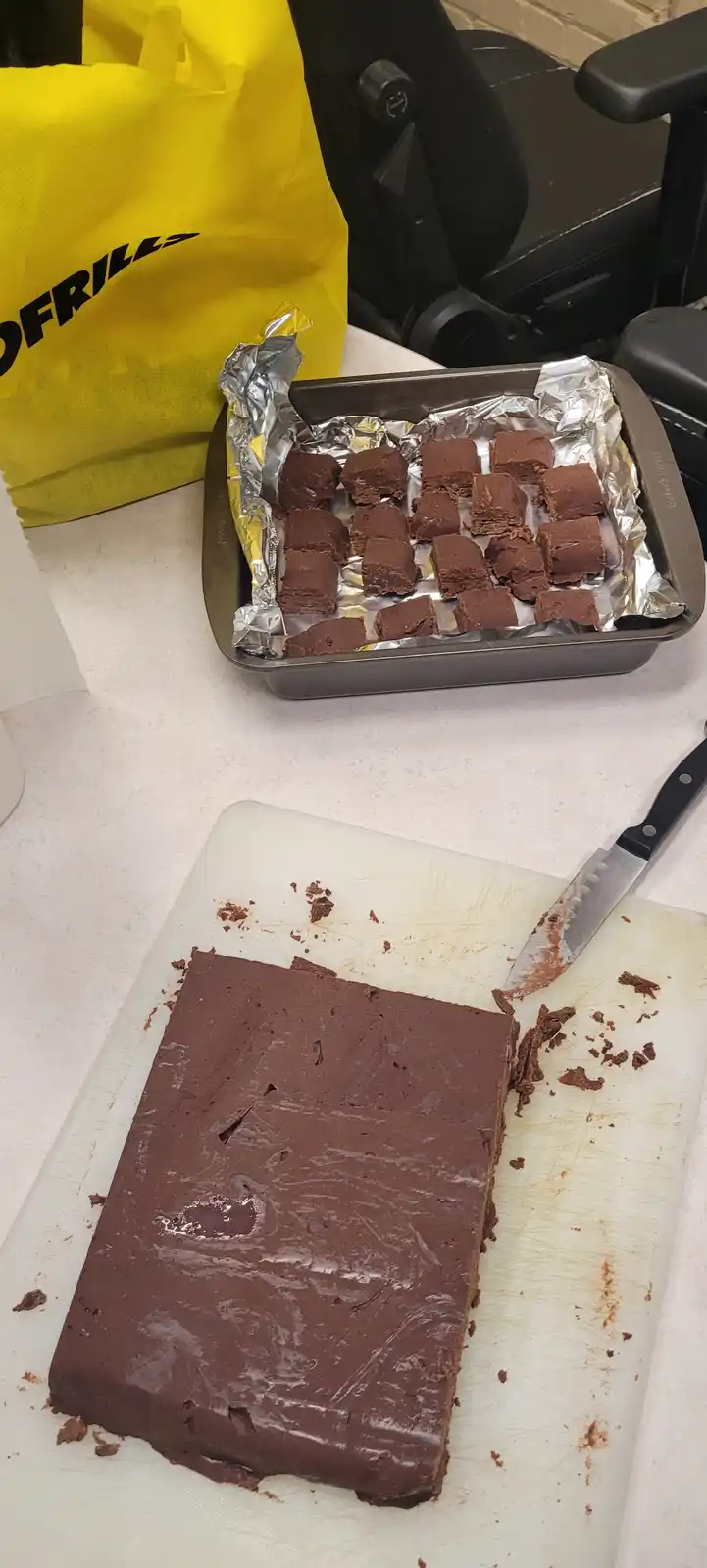A kitchen counter shows a large, freshly cut block of chocolate fudge on a white cutting board with a knife beside it, while…