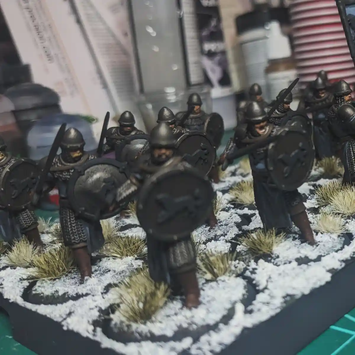 A photo of painted miniature fantasy soldiers in dark armor, holding shields with a horse symbol and swords, standing on a…
