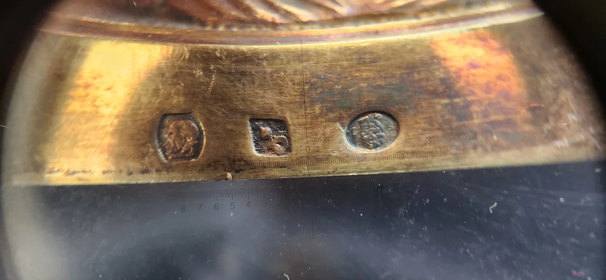A close-up photo shows three small, worn coins embedded in a brass ruler with measurement markings. The ruler is placed on a…
