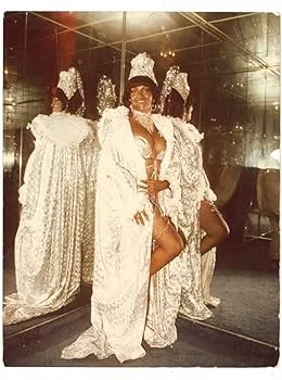 A woman in a lavish, white, bejeweled gown and crown poses confidently in front of a mirror, her reflection showing multiple…