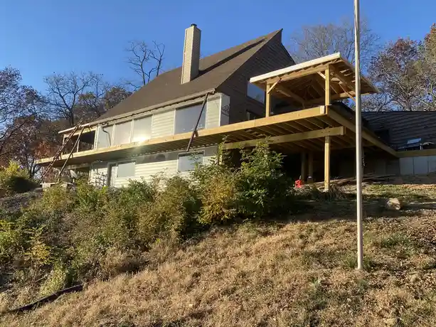A house under construction with exposed wooden framing for a large deck and porch, set against a backdrop of trees and a…